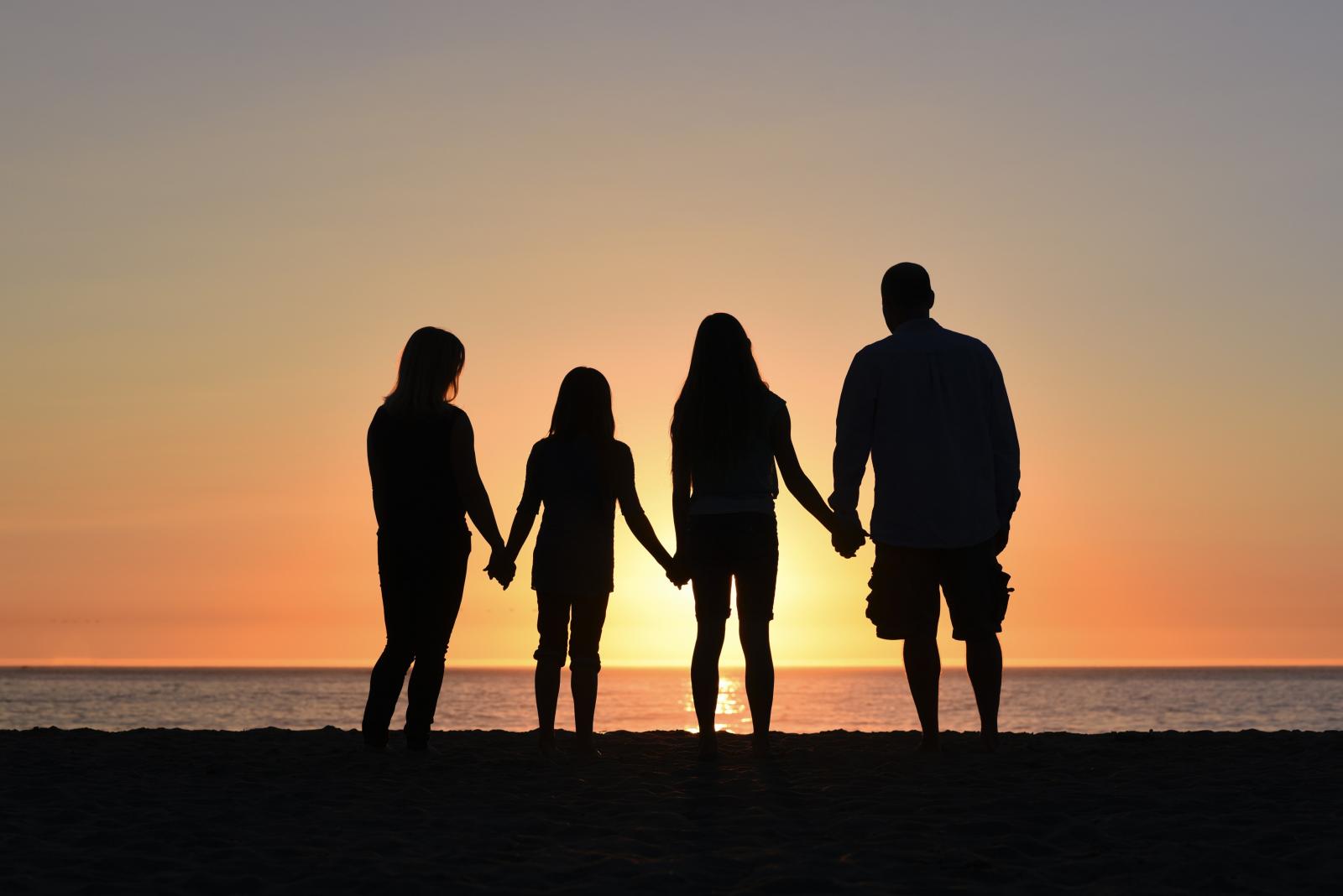 Quattro persone viste di spalle (una donna, un uomo, due ragazze) si tengono per mano davanti al mare e al tramonto