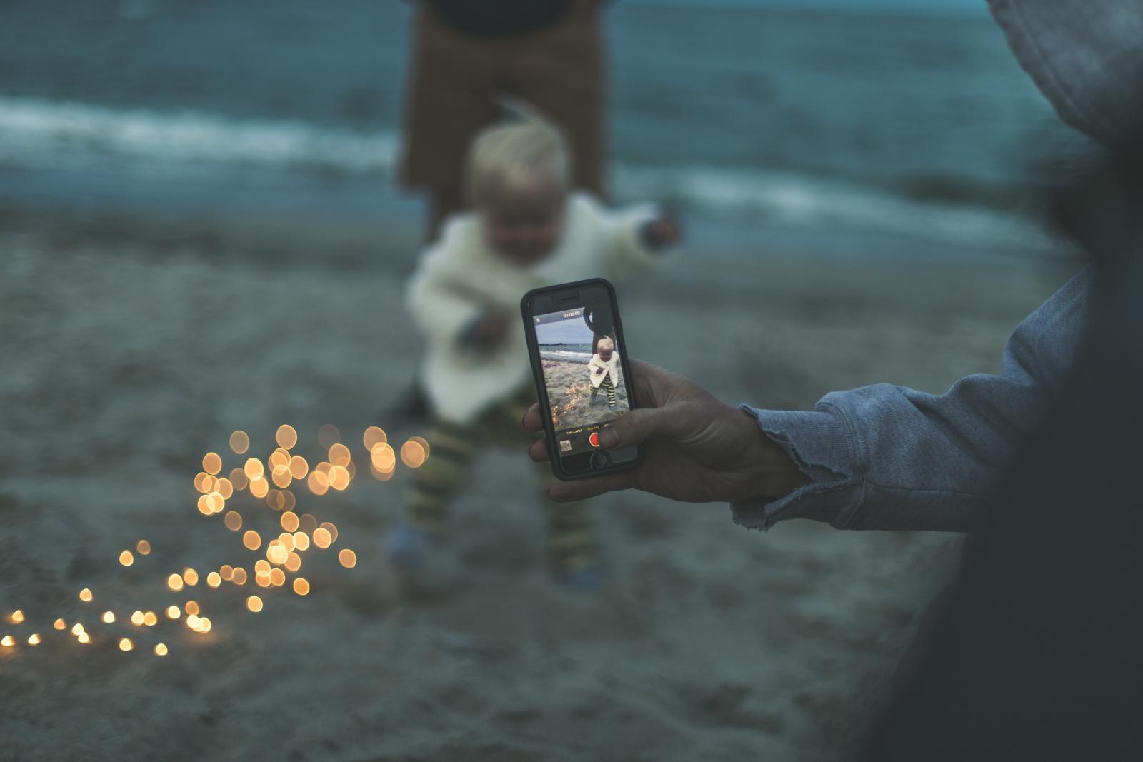 Una bambina piccola sulla spiaggia viene fotograta con il telefono.