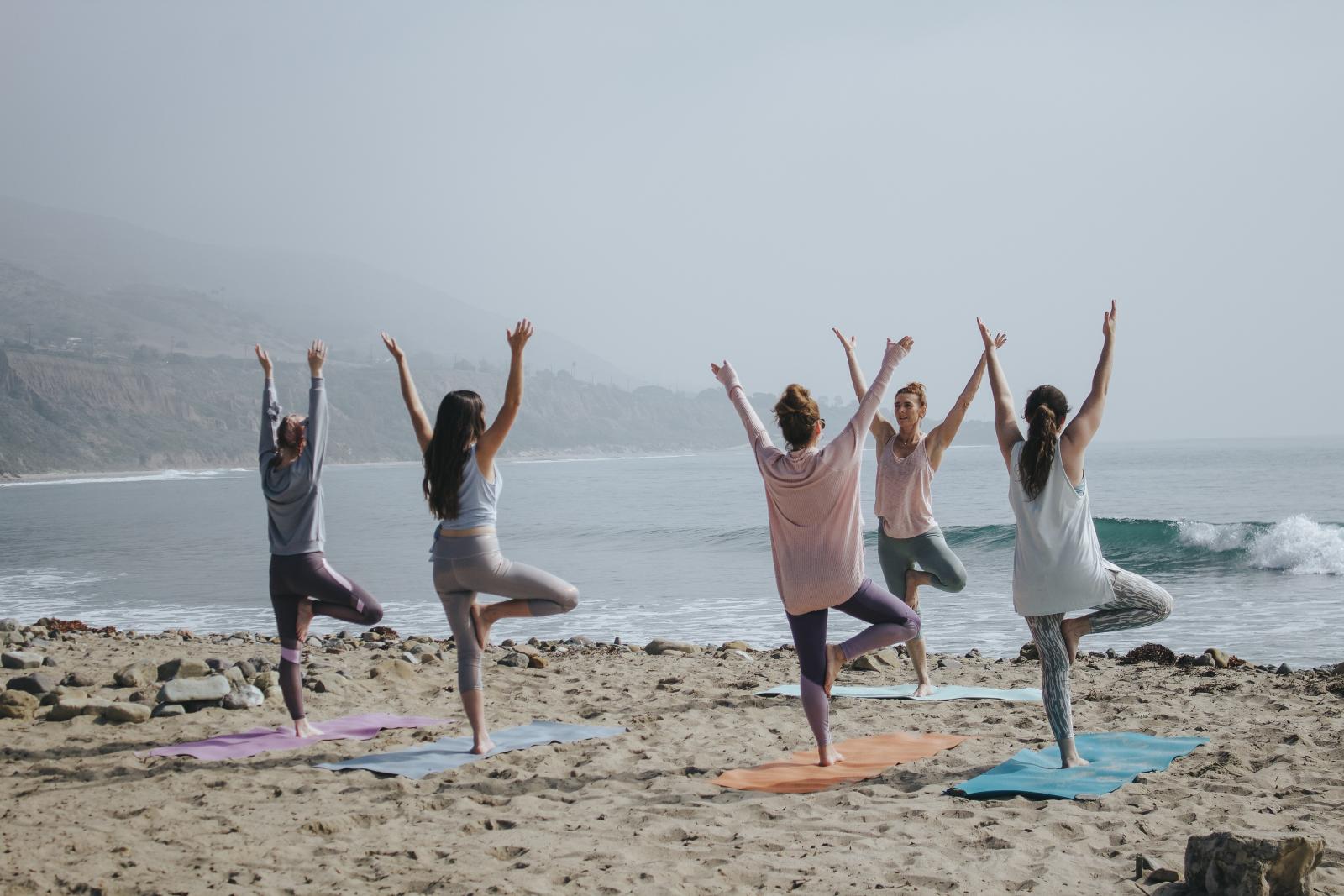 Alcune ragazze che praticano yoga sulla spiaggia, con braccia in alto, sollevate su un piede