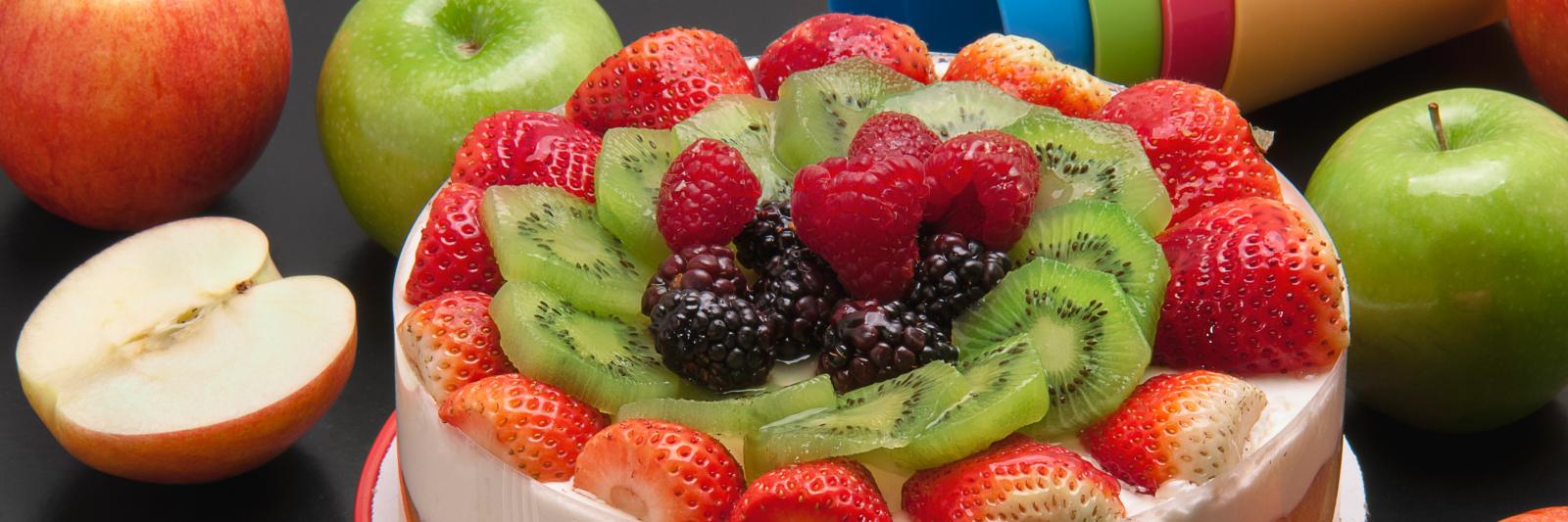 Cheescake ricoperta di frutta fresca; accanto mele, posate e bicchieri di plastica