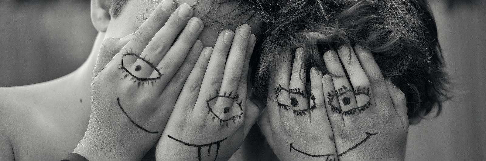 Bambini che si coprono il volto con le mani, sulle mani il disegno di due facce spiritose