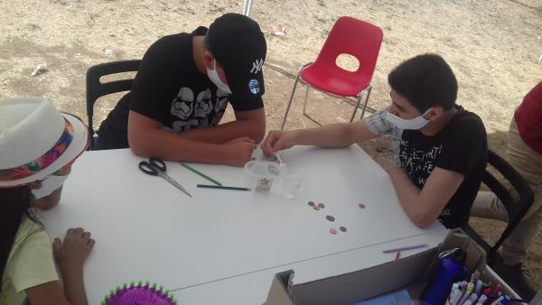 Tre ragazzi seduti ad un tavolo che stanno realizzando un esperimento, sul tavolo alcune monete, forbici, bastoncini.