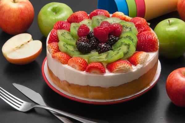 Cheescake ricoperta di frutta fresca; accanto mele, posate e bicchieri di plastica