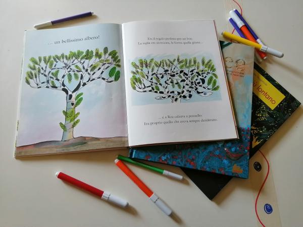 Libro illustrato aperto sul tavolo (c'è disegnato un albero). Accanto altri libri e pennarelli colorati.